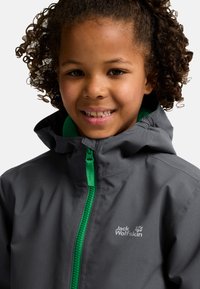Enfant aux cheveux bouclés portant une veste grise Jack Wolfskin avec une fermeture éclair verte, souriant à la caméra.