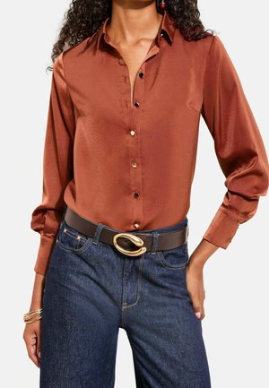 Kvinde iført en skinnende rustfarvet langærmet skjorte med knapper, mørkeblå jeans og et sort bælte med en stor guldlås.