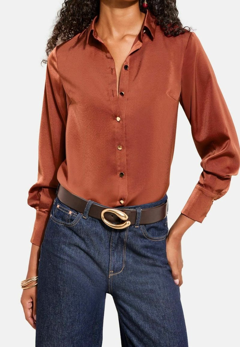 Femme portant une chemise à manches longues brillante couleur rouille, un jean bleu foncé et une ceinture noire avec une grosse boucle dorée.