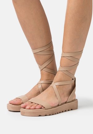Beige flache Sandalen mit dicken Sohlen und überkreuzten Knöchelriemen, getragen auf baren Füßen und Unterschenkeln vor einem weißen Hintergrund.