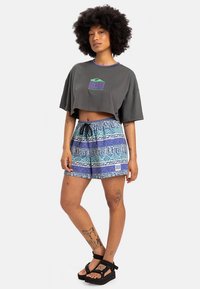 Grå kort t-shirt med lila kant och en grön logotyp. Blå kortbyxor med geometriska mönster och snörning i midjan. Svarta platåsandaler.