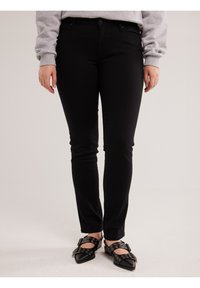 Persona vistiendo pantalones ajustados negros, zapatos negros puntiagudos con hebillas y ojales, y una camiseta de manga larga gris claro, de pie sobre un suelo blanco.