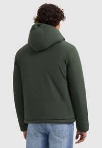 Groene capuchonjas met een gladde, matte textuur, met een relaxte pasvorm en elastische manchetten. De achterkant toont minimale details en naden.