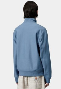 Sudadera azul de cuello alto y puños acanalados, fabricada en un material suave; presenta una pequeña etiqueta con el logo en la parte inferior de la espalda.