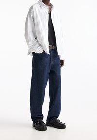 Homme portant un jean bleu foncé ample, une ceinture noire, une chemise noire, une chemise à rayures blanches boutonnée et des mocassins noirs, debout les mains dans les poches.