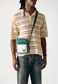Borsa a tracolla con un corpo crema, flap verde, accenti blu navy e un logo prominente. Chi indossa la borsa ha una maglietta in maglia testurizzata e jeans.