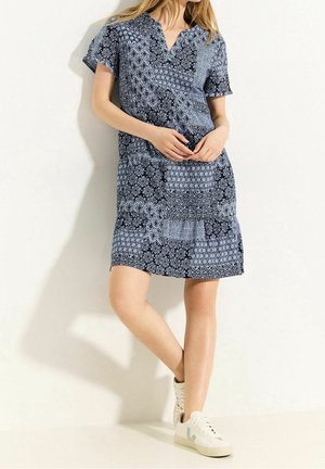 Femme portant une robe bleu marine à manches courtes, longueur genou, avec des motifs floraux et géométriques blancs, debout les jambes croisées, baskets blanches.