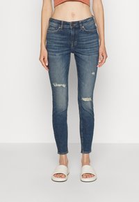 Vaqueros de mezclilla azul ajustados con parches desgastados, cintura alta y diseño de cinco bolsillos. Llevados con sandalias blancas tipo slide.