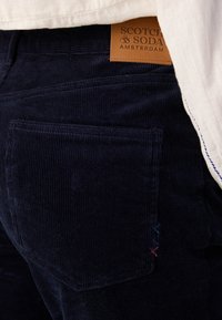 Marineblaue Corduroyhosen mit einem Lederetikett am Bund und einer Gesäßtasche mit dekorativer roten und blauen Stickerei.