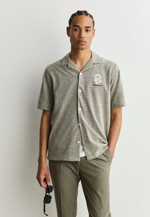 Joven con el pelo trenzado, vistiendo una camisa gris de manga corta abotonada y pantalones verdes, sosteniendo unas gafas de sol negras en su mano izquierda.