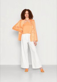Blazer laranja riscado sobre uma camiseta laranja, combinado com calças largas brancas e sandálias de salto laranja. Tecidos suaves e ajuste relaxado.