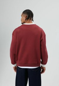 Bordeauxrode sweatshirt met een losse pasvorm, brede manchetten en een witte halslijn. De stof lijkt zacht en heeft een casual ontwerp, geschikt voor ontspannen draagmomenten.