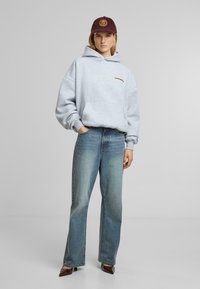 Felpa grigia con cappuccio e tasca frontale, con testo rosso, abbinata a jeans a gamba larga blu scoloriti e un cappellino bordeaux con logo texturizzato.