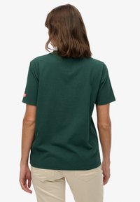 Donkergroene katoenen T-shirt met korte mouwen, ronde hals en een klein rood logoplaatje op de linkermouw. Zachte textuur en standaard pasvorm.