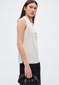 Ανοιχτό μπεζ αμάνικο τοπ με ντραπέ λαιμόκοψη, απαλή υφή και χαλαρή εφαρμογή, συνδυασμένο με μια μαύρη δερμάτινη τσάντα χειρός.