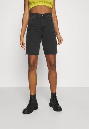 Personne portant un short en jean noir taille haute avec des ourlets bruts, des bottes noires à lacets et un top court jaune, debout devant un fond uni.