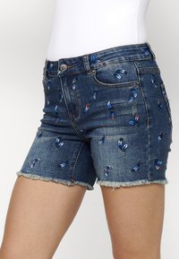 Denim šorti ar izbalinātu zilu krāsu, kas rotāti ar izšūtām zīda rakstiem z blue, sarkanā un baltā krāsā. Saplēsts malu dizains un piecu kabatu stilā.