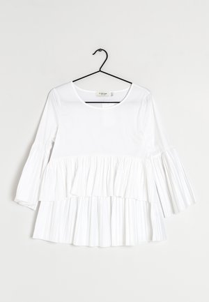 Biała bluzka z długim rękawem z warstwami plisowanych falban na rękawach i na dole, wisząca na czarnym wieszaku na jednolitym tle.