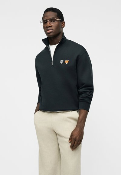 Άνδρας που φοράει διάφανα γυαλιά, μαύρο φούτερ με φερμουάρ μέχρι τη μέση με μπαλώματα λύκου και αλεπούς, και ανοιχτό μπεζ παντελόνι, στέκεται μπροστά σε απλό φόντο.