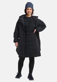 Giacca nera isolante con cappuccio, gonna abbottonata e leggings neri aderenti. Berretto blu a coste e scarpe da ginnastica nere.