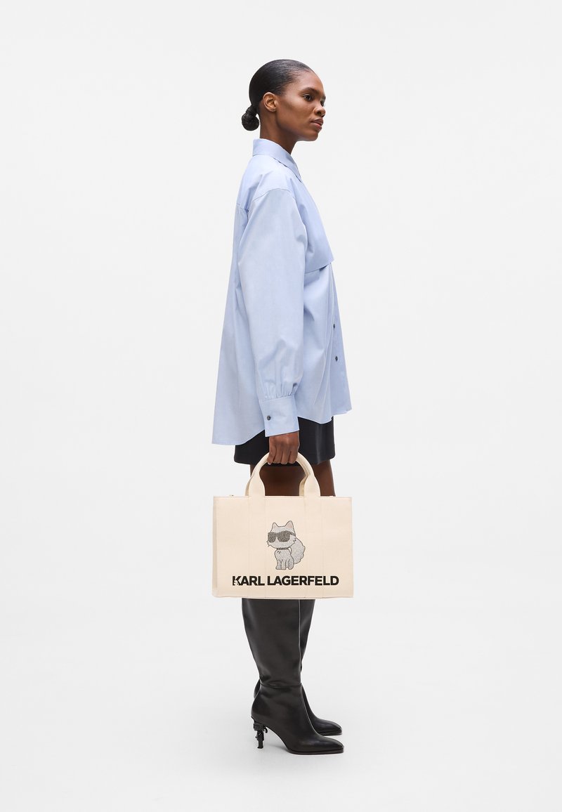 Chemise bleu clair à boutons, mini-jupe noire, bottes hautes noires à talons, et sac fourre-tout crème avec un graphique et le texte "KARL LAGERFELD."