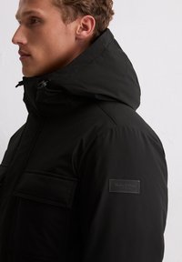 Zwarte gewatteerde jas met een hoge kraag, capuchon met trekkoord en twee zakken aan de voorkant. Subtiele logopatch op de mouw. Glad oppervlak.