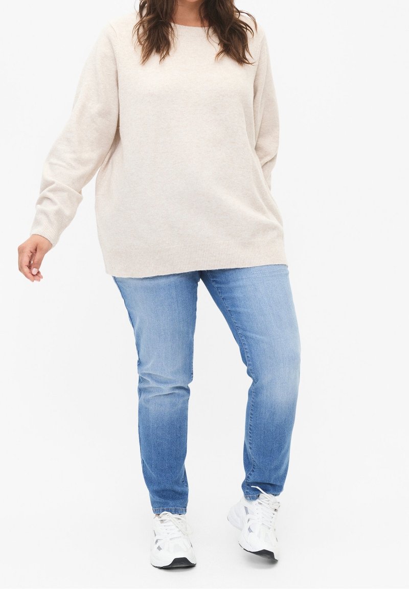 Maglione beige over size con maniche lunghe, abbinato a jeans azzurri e sneakers bianche, su sfondo bianco semplice.