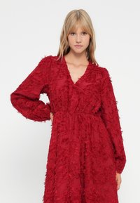 Junge Frau mit blonden Haaren, die ein strukturiertes, tiefrotes langärmliges V-Ausschnitt-Kleid trägt und mit einer Hand an der Hüfte vor einem weißen Hintergrund posiert.