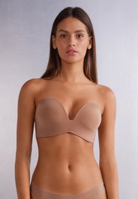 Bezešvá podprsenka v béžové barvě, hladká tkanina, konturovaný tvar a bezšvový design. Má zakřivený výstřih a minimalistický styl.