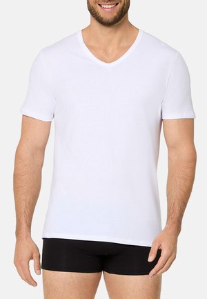 Mann lächelnd trägt ein schlichtes weißes kurzärmeliges V-Ausschnitt-Shirt und schwarze, eng anliegende Shorts vor hellem Hintergrund.