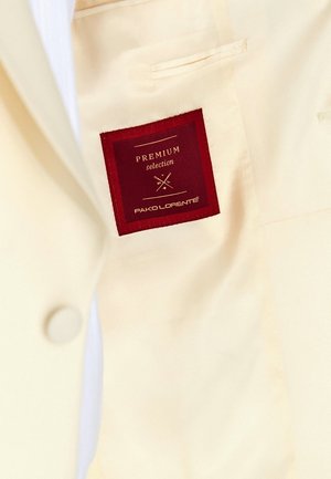 Wnętrze kremowego garnituru z czerwoną metką z napisem "PREMIUM selection" i "PAKO LORENTE". Gładka tkanina z widocznym detalem guzika.