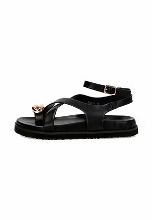Sandalia plana negra con tiras cruzadas, hebilla ajustable en el tobillo y pequeño detalle dorado en forma de corazón en la lazada delantera del dedo.