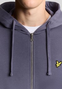 Grauer Kapuzenpullover mit Reißverschluss aus weichem Stoff, mit Kängurutasche, Kordelzugkapuze und gelbem gesticktem Logo auf der linken Brust.