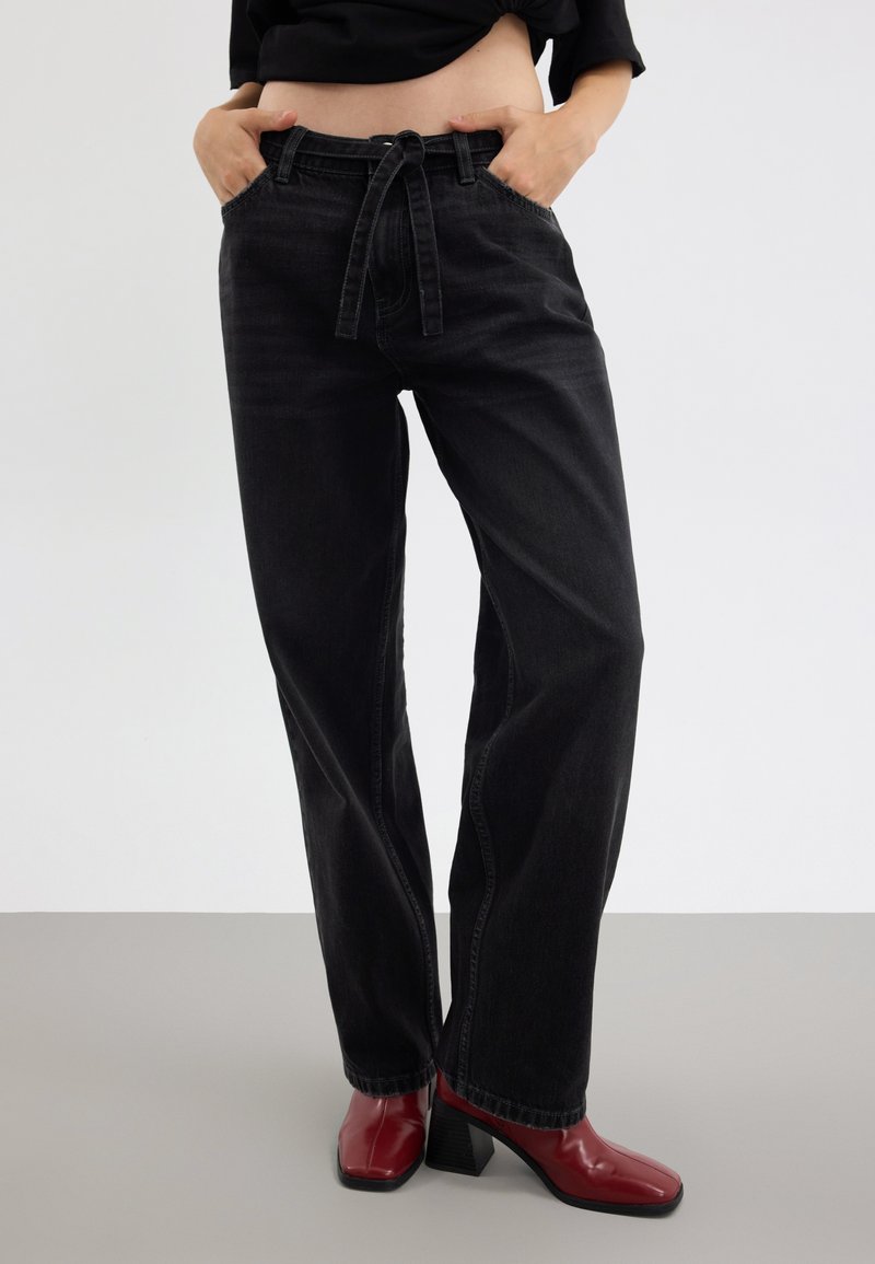 Črne jeans hlače ohlapnega kroja z vrvico v pasu, ki imajo sprednje žepe in značilno šivanje na nogah, v kombinaciji z rdečimi visokimi čevlji.