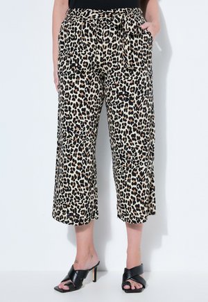 Frau trägt kurze, weit geschnittene Leopardenmusterhose mit seitlichen Taschen und schwarzen Sandalen mit Absatz, steht vor weißem Hintergrund.