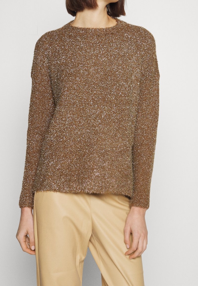 Donna con un maglione metallico marrone e peloso a maniche lunghe abbinato a pantaloni beige, rivolta leggermente di lato su uno sfondo bianco.