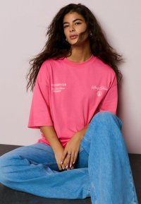 Roza bombažna prevelika majica z belim napisom spredaj, kombinirana s svetlo modrimi širokimi kavbojkami. Model sedi z rokami nameščenimi na kolenih.