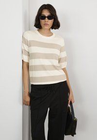 Lichtgewicht, gestreept gebreid top in beige en crème met een ronde hals en korte mouwen, gecombineerd met zwarte broek en een handtas.