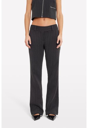 Femme portant un pantalon noir à fines rayures taille haute associé à un gilet court assorti et des chaussures noires à bout pointu.