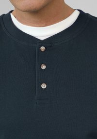 Marineblauw henley-shirt met een textuurstof, een ronde halslijn en drie lichtgekleurde knopen aan de voorkant. Witte onderhemd zichtbaar.
