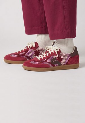 Baskets rouges et violettes à motifs avec des lacets blancs, portées avec des chaussettes blanches et un pantalon rouge court sur un fond neutre.