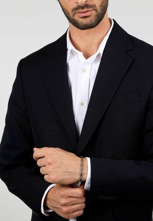 Bracciale argentato con anelli rettangolari, indossato su un polso. Il portatore indossa un blazer blu navy sopra una camicia bianca.
