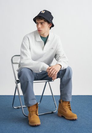 Giovane uomo che indossa un cappello a secchiello nero, una maglia a maniche lunghe grigio chiaro, pantaloni blu e stivali da lavoro color sabbia, seduto su una sedia pieghevole in metallo su un tappeto blu.