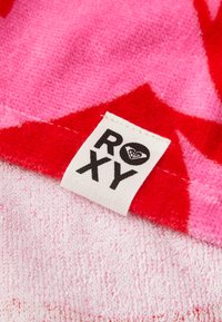 Toalla de la marca Roxy en un vibrante color rosa y rojo con una textura suave, que presenta un logotipo de corazón en la etiqueta y patrones abstractos.