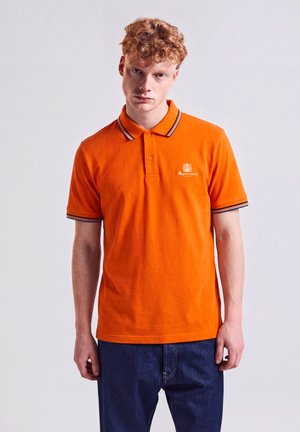 Junger Mann mit lockigem roten Haar, der ein orangefarbenes Polohemd und dunkelblaue Jeans trägt und vor einem einfarbigen hellen Hintergrund steht.