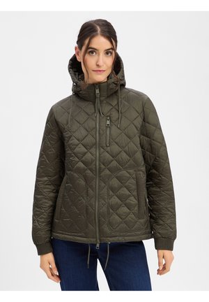 Woman wearing a dark green quilted hooded jacket with zipper front and pockets, paired with dark blue jeans, standing against white background.