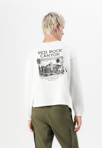 Biela dlhá rukávová košeľa s čiernou grafikou znázorňujúcou Red Rock Canyon, so textom a obrazmi púšte. Doplnená olivovozelenými nohavicami.