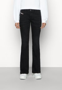 Persona con jeans de bota ancha negros, sudadera blanca y zapatillas de plataforma blancas, de pie contra un fondo claro y liso.