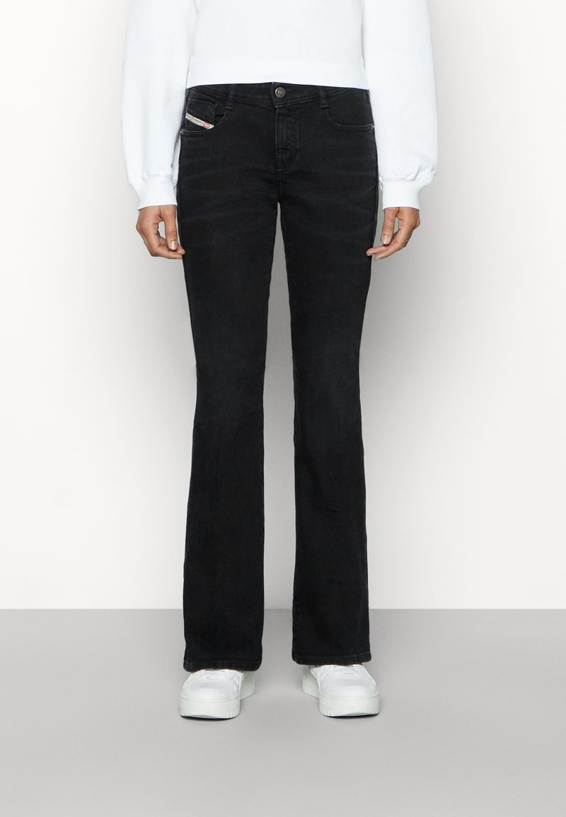 Persona con jeans de bota ancha negros, sudadera blanca y zapatillas de plataforma blancas, de pie contra un fondo claro y liso.