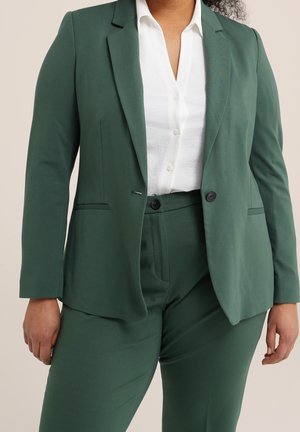 Femme portant un costume vert foncé avec un blazer à un seul bouton et un pantalon assorti, accompagné d'une chemise blanche à boutons.